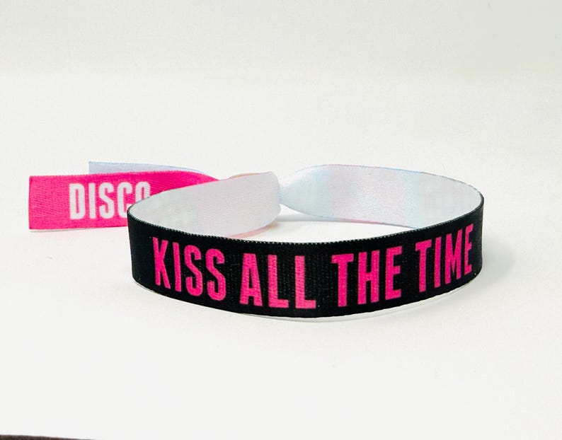 Może przedstawiać: Czarna opaska na nadgarstek z r&oacute;żowym napisem "KISS ALL THE TIME". Widoczna jest r&oacute;wnież oddzielna r&oacute;żowa etykieta z napisem "DISCO". Opaska wykonana jest z tkanego materiału.