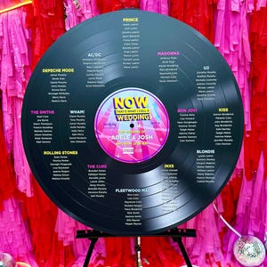 Può includere: Un disco in vinile nero con un'etichetta bianca che dice "Now That's What I Call a Wedding" e presenta i nomi Adele & Josh. Il disco è circondato da un elenco di artisti musicali e i loro nomi, tra cui Prince, Madonna, U2, Bon Jovi, Kiss, Blondie, The Smiths, Wham!, Depeche Mode, AC/DC, The Cure, Fleetwood Mac, INXS e Rolling Stones.