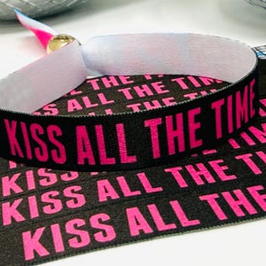 Może przedstawiać: Czarna opaska na nadgarstek z tkaniny z napisem "KISS ALL THE TIME" w jaskrawym r&oacute;żu. Opaska ma białe wnętrze i złote zapięcie. W tle widocznych jest kilka podobnych opasek.