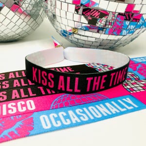 Może przedstawiać: Czarna opaska na nadgarstek z r&oacute;żowym napisem "KISS ALL THE TIME". Opaska leży na r&oacute;żowo-niebieskiej tkaninie we wzory ze słowami "DISCO" i "OCCASIONALLY". Dwie kule dyskotekowe w tle.