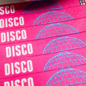 Może przedstawiać: Zbliżenie na r&oacute;żowe opaski na nadgarstki z białym napisem "DISCO" i niebieską grafiką kuli dyskotekowej. Opaski są ułożone w stos, a w tle widoczna jest lustrzana kula dyskotekowa. Opaski są prawdopodobnie przeznaczone na imprezę.