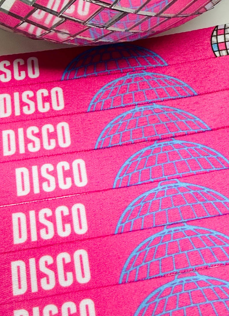 Może przedstawiać: Zbliżenie na r&oacute;żowe opaski na nadgarstki z białym napisem "DISCO" i niebieską grafiką kuli dyskotekowej. Opaski są ułożone w stos, a w tle widoczna jest lustrzana kula dyskotekowa. Opaski są prawdopodobnie przeznaczone na imprezę.