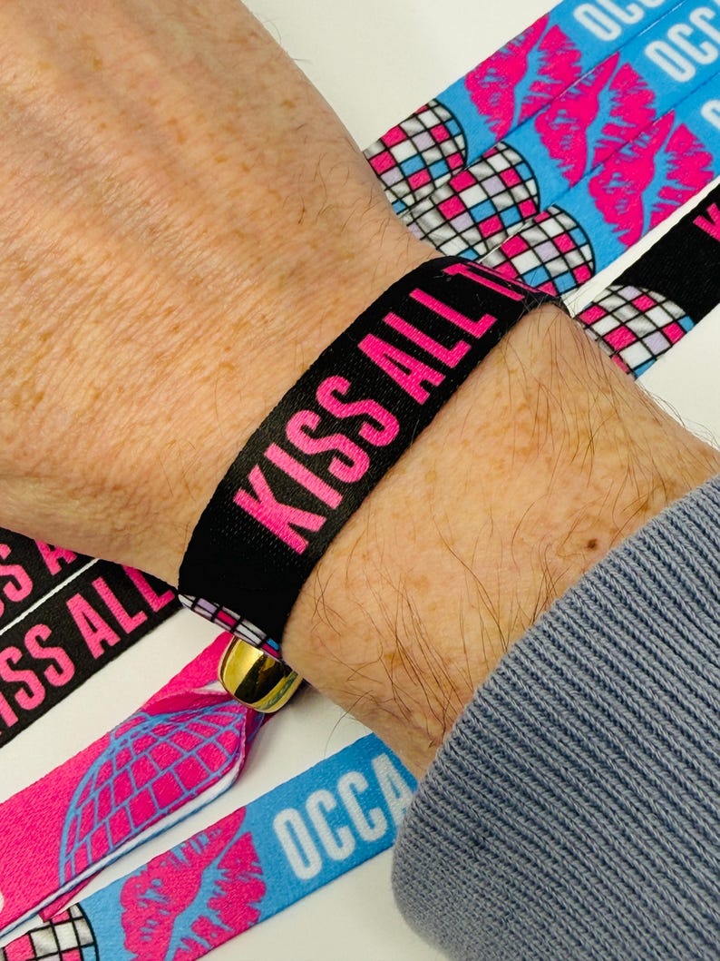 Może przedstawiać: Czarna opaska na nadgarstek z tkaniny z napisem "KISS ALL" w jaskrawym r&oacute;żu. Widoczne są dodatkowe opaski z grafiką kuli dyskotekowej i ust. Opaska jest zabezpieczona złotym zapięciem. Og&oacute;lny motyw związany jest z wydarzeniami lub imprezami.