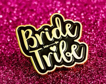 May include: Gold-colored pin with black lettering that reads "Bride Tribe" with a small arrow and heart in the center. The pin is on a pink sparkly background.