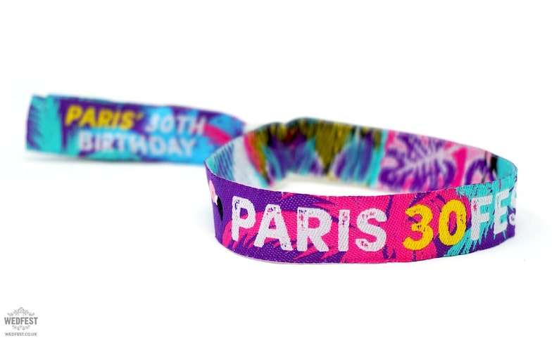 May include: A colorful wristband with the text "Paris 30th Festival" in white letters on a pink background. The wristband also has a tropical print with blue, green, yellow, and pink colors.