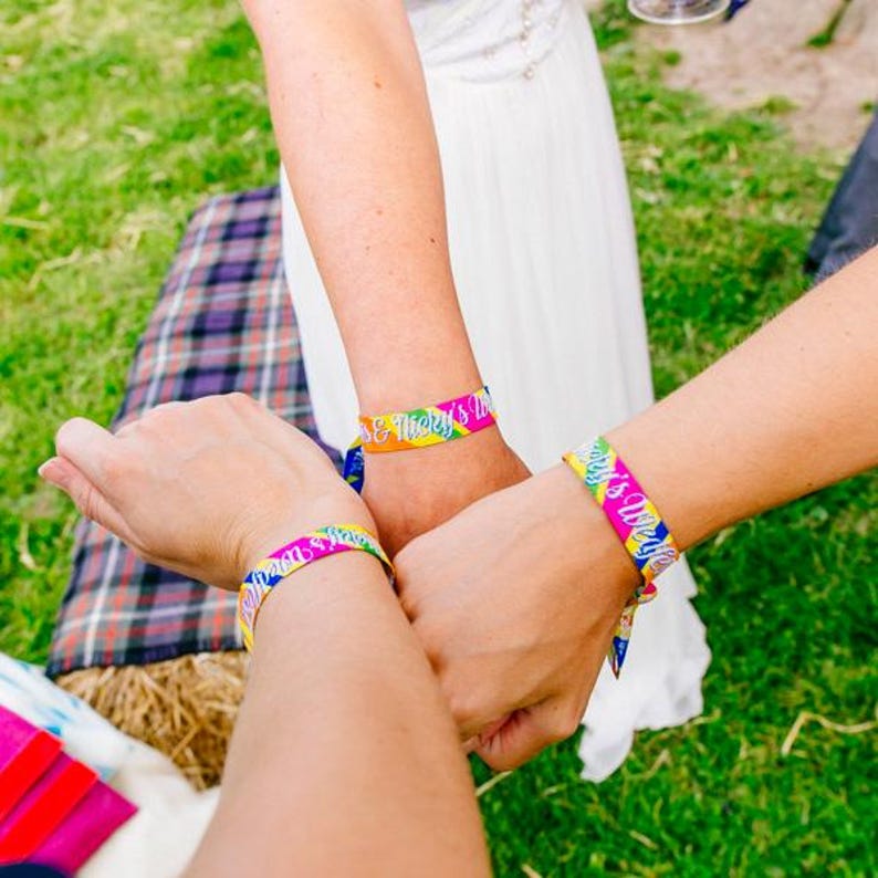May include: Three people wearing colorful wristbands with the text "[Name] & [Name]'s Wedding" and "[Name]'s Wedding".