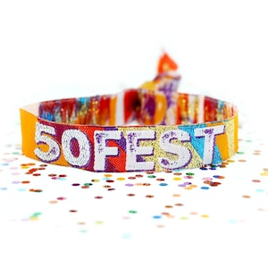 Może przedstawiać: Kolorowa opaska na nadgarstek z tkaniny z białym napisem "50FEST". Opaska ma żywy wzór w kolorze pomarańczowym, czerwonym, fioletowym i złotym. Tło jest białe z rozrzuconym konfetti.