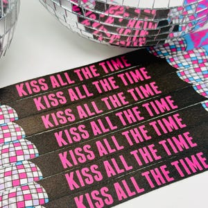 Może przedstawiać: Zbliżenie czarnych opasek na nadgarstki z napisem "KISS ALL THE TIME" w jaskrawym r&oacute;żu. Opaski są ułożone w pobliżu kuli dyskotekowej z lustrzanymi kafelkami. Kula dyskotekowa odbija r&oacute;żowe i niebieskie odcienie, tworząc żywy obraz o tematyce imprezowej.