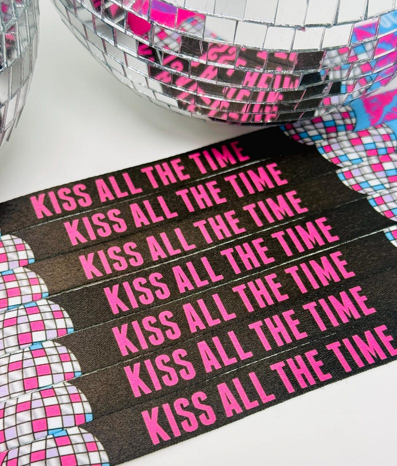 Może przedstawiać: Zbliżenie czarnych opasek na nadgarstki z napisem "KISS ALL THE TIME" w jaskrawym r&oacute;żu. Opaski są ułożone w pobliżu kuli dyskotekowej z lustrzanymi kafelkami. Kula dyskotekowa odbija r&oacute;żowe i niebieskie odcienie, tworząc żywy obraz o tematyce imprezowej.