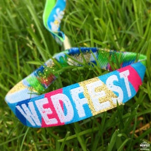 May include: A colorful wristband with the word "WEDFEST" printed in white letters with gold glitter accents. The wristband is made of fabric and has a blue, pink, and white striped pattern.