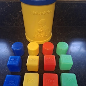 Może przedstawiać: Zestaw zabawek Fisher-Price w stylu vintage. Zawiera żółty cylinder z niebieską pokrywą i różne kolorowe klocki. Zestaw zawiera niebieskie, żółte, czerwone i zielone klocki w kształcie cylindrycznym i sześciennym. Cylinder ma wytłoczoną postać dziecka.