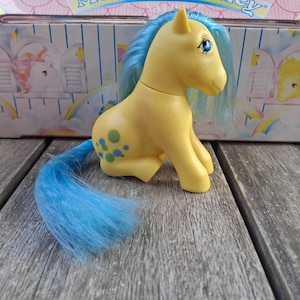 Puede incluir: Un poni de juguete amarillo con pelo y cola azules, y manchas azules y verdes en el costado. El poni está sentado sobre una superficie de madera, con una caja en el fondo con una ilustración de poni similar y un diseño de arcoíris.