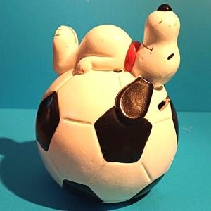 May include: A white ceramic Snoopy figure resting on a football. Snoopy has a black nose and ears, and a red collar. The football is white with black pentagons. A decorative item.