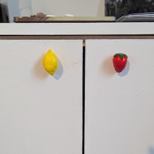 Könnte beinhalten: Zwei dekorative Schrankknöpfe: eine gelbe Zitrone und eine rote Erdbeere mit grünem Oberteil. Sie sind an einer weißen Schranktür befestigt.