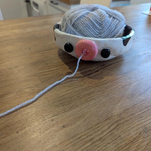 Puede incluir: Un cuenco de cerámica hecho a mano para lana con diseño de vaca. El cuenco es blanco con manchas negras y una guía de hilo rosa. Una madeja de lana gris se encuentra dentro del cuenco, con el hilo saliendo. El cuenco está sobre una mesa de madera.