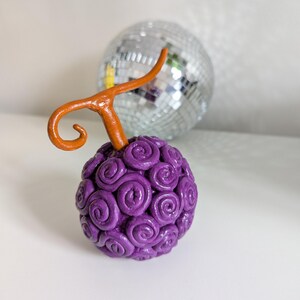 Op de afbeelding: Een handgemaakt beeldje van de "Gum-Gum Fruit" uit de anime-serie One Piece. De vrucht is een bol met een swirl-patroon en een enkele oranje steel.