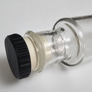 Puede incluir: Una botella de vidrio transparente con una tapa negra. La botella tiene un cuello blanco y un diseño decorativo grabado en el vidrio. La tapa está hecha de un material oscuro y acanalado. La botella es probablemente para almacenar líquidos.