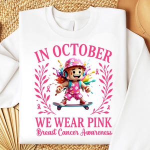 May include: White sweatshirt with a graphic promoting Breast Cancer Awareness. The design features a cartoon character on a skateboard, surrounded by pink ribbons and the text "In October We Wear Pink".