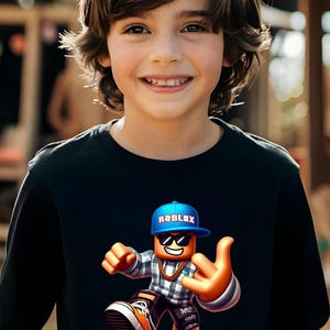 以下が含まれることがあります： 笑顔の子供が、Robloxのキャラクターのカラフルなグラフィックが描かれた黒い長袖シャツを着ています。キャラクターは「ROBLOX」の文字が入った青いキャップ、サングラス、金のチェーンを身につけています。キャラクターは手のジェスチャーをしています。