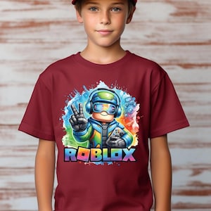 K&ouml;nnte beinhalten: Ein weinrotes T-Shirt mit einer farbenfrohen Grafik eines Roblox-Charakters mit Kopfh&ouml;rern und einem Controller, der ein Friedenszeichen zeigt. Das Wort "ROBLOX" ist in Regenbogenfarben dargestellt. Die Person im Bild tr&auml;gt eine weinrote Baseballkappe.