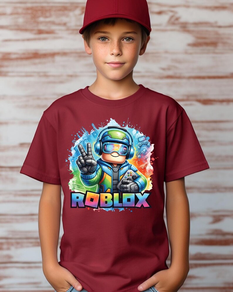 K&ouml;nnte beinhalten: Ein weinrotes T-Shirt mit einer farbenfrohen Grafik eines Roblox-Charakters mit Kopfh&ouml;rern und einem Controller, der ein Friedenszeichen zeigt. Das Wort "ROBLOX" ist in Regenbogenfarben dargestellt. Die Person im Bild tr&auml;gt eine weinrote Baseballkappe.