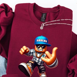 以下が含まれることがあります： ロブロックスのキャラクターグラフィックが特徴のマルーンスウェットシャツ。キャラクターは、ロブロックスのロゴが入った青い帽子、サングラス、金のチェーンを着用しています。スウェットシャツはクルーネックで、シルバーのネックレスが付いています。