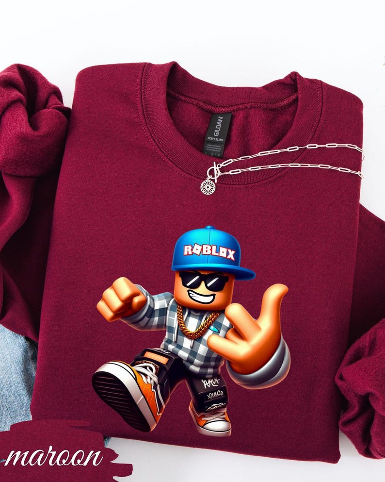 以下が含まれることがあります： ロブロックスのキャラクターグラフィックが特徴のマルーンスウェットシャツ。キャラクターは、ロブロックスのロゴが入った青い帽子、サングラス、金のチェーンを着用しています。スウェットシャツはクルーネックで、シルバーのネックレスが付いています。