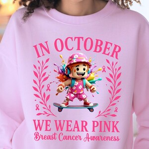 May include: Pale pink sweatshirt with the message "IN OCTOBER WE WEAR PINK Breast Cancer Awareness." The design includes a cartoon figure on a skateboard, surrounded by pink ribbons and colourful paint.