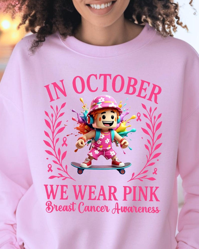 May include: Pale pink sweatshirt with the message "IN OCTOBER WE WEAR PINK Breast Cancer Awareness." The design includes a cartoon figure on a skateboard, surrounded by pink ribbons and colourful paint.