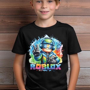 K&ouml;nnte beinhalten: Schwarzes T-Shirt mit einer farbenfrohen Grafik eines Charakters, der ein Headset tr&auml;gt und einen Gamecontroller h&auml;lt. Das Wort "ROBLOX" wird in Regenbogenfarben unterhalb der Figur angezeigt. Das Bild hat einen Aquarelleffekt.