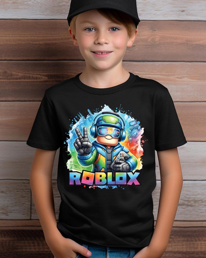 K&ouml;nnte beinhalten: Schwarzes T-Shirt mit einer farbenfrohen Grafik eines Charakters, der ein Headset tr&auml;gt und einen Gamecontroller h&auml;lt. Das Wort "ROBLOX" wird in Regenbogenfarben unterhalb der Figur angezeigt. Das Bild hat einen Aquarelleffekt.