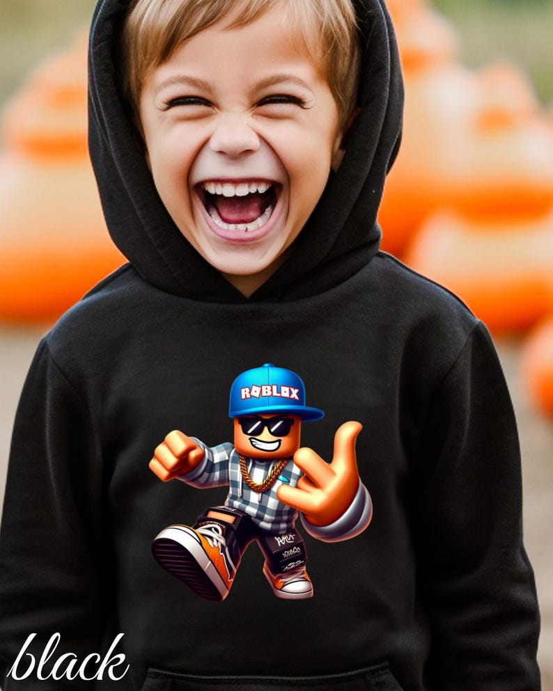 以下が含まれることがあります： 黒いフード付きスウェットシャツ。カラフルなRobloxキャラクターのグラフィックが特徴です。キャラクターは青いキャップ、サングラス、金のチェーンを身に着けています。「black」という単語が画像の下部にイタリック体で書かれています。