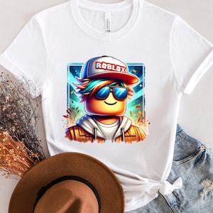 May include: A white t-shirt with a colorful cartoon image of a Roblox character wearing sunglasses and a hat that says 'Roblox' on it. The character is against a blue and purple background with stars and confetti.