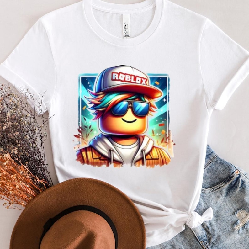 May include: A white t-shirt with a colorful cartoon image of a Roblox character wearing sunglasses and a hat that says 'Roblox' on it. The character is against a blue and purple background with stars and confetti.