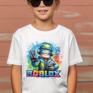 K&ouml;nnte beinhalten: Wei&szlig;es T-Shirt mit einem farbenfrohen Roblox-Grafikdesign. Das Design zeigt eine Figur mit Headset und Gamecontroller, sowie das Wort "ROBLOX" in Regenbogenfarben. Das T-Shirt wird von einer Person mit Sonnenbrille und schwarzer Kappe getragen.
