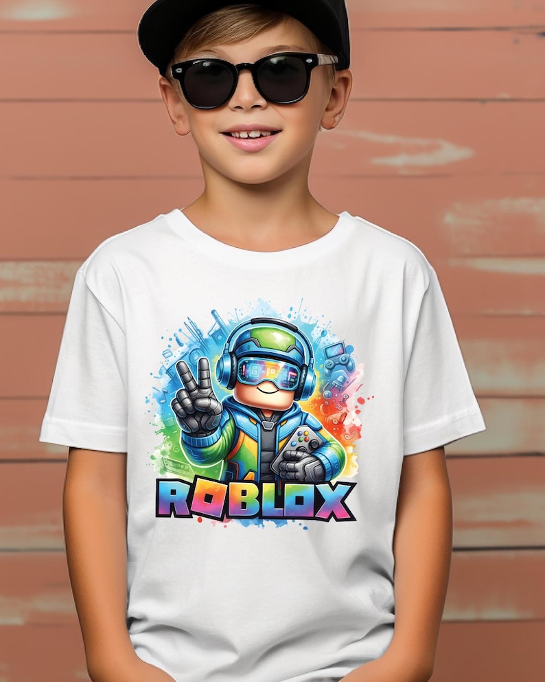 K&ouml;nnte beinhalten: Wei&szlig;es T-Shirt mit einem farbenfrohen Roblox-Grafikdesign. Das Design zeigt eine Figur mit Headset und Gamecontroller, sowie das Wort "ROBLOX" in Regenbogenfarben. Das T-Shirt wird von einer Person mit Sonnenbrille und schwarzer Kappe getragen.