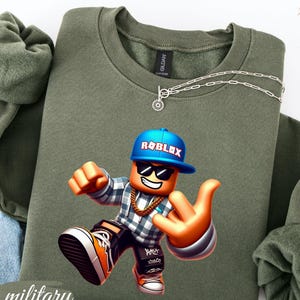 以下が含まれることがあります： ミリタリーグリーンのスウェットシャツ。カラフルなRobloxキャラクターのグラフィックが特徴です。キャラクターは青いキャップ、サングラス、金のチェーンを身に着けています。スウェットシャツはクルーネックで、シルバーのネックレスが付いています。「military green」の文字が左下に印刷されています。