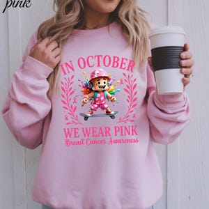 May include: Pale pink sweatshirt with the message "IN OCTOBER WE WEAR PINK Breast Cancer Awareness". The design includes a cartoon figure on a skateboard, pink ribbons, and a floral wreath, promoting breast cancer awareness.