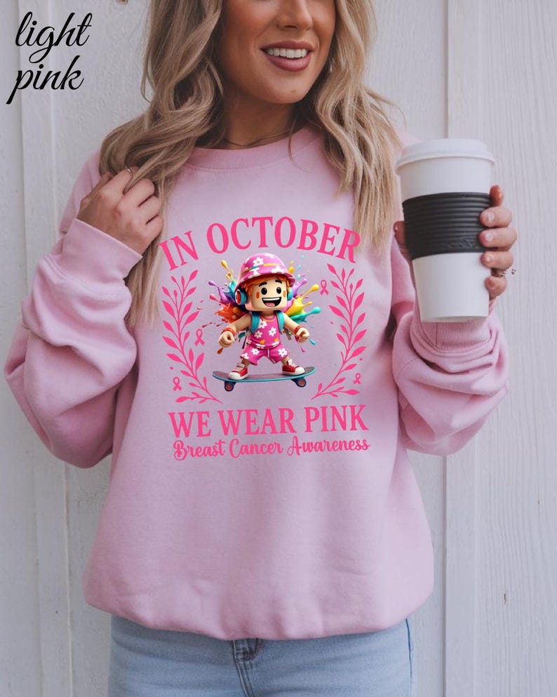May include: Pale pink sweatshirt with the message "IN OCTOBER WE WEAR PINK Breast Cancer Awareness". The design includes a cartoon figure on a skateboard, pink ribbons, and a floral wreath, promoting breast cancer awareness.