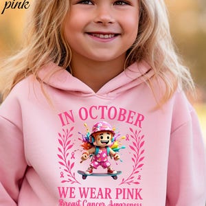 May include: Pale pink hoodie with the text "IN OCTOBER WE WEAR PINK Breast Cancer Awareness". The hoodie features a graphic of a girl on a skateboard with colourful accents and pink ribbons. The words "light pink" are in the upper left corner.