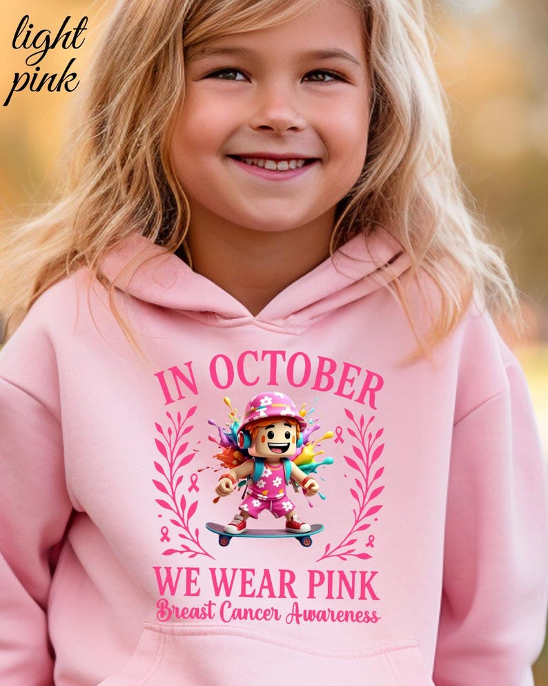May include: Pale pink hoodie with the text "IN OCTOBER WE WEAR PINK Breast Cancer Awareness". The hoodie features a graphic of a girl on a skateboard with colourful accents and pink ribbons. The words "light pink" are in the upper left corner.