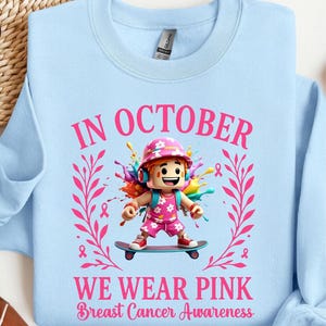 May include: Pale blue sweatshirt with the text "IN OCTOBER WE WEAR PINK Breast Cancer Awareness." The design features a cartoon character on a skateboard, surrounded by pink ribbons and floral elements. The sweatshirt is light blue.