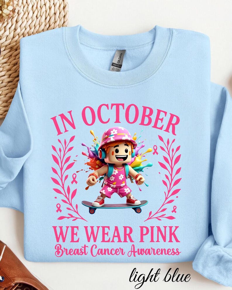 May include: Pale blue sweatshirt with the text "IN OCTOBER WE WEAR PINK Breast Cancer Awareness." The design features a cartoon character on a skateboard, surrounded by pink ribbons and floral elements. The sweatshirt is light blue.