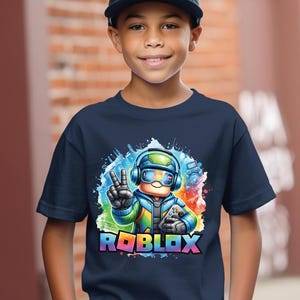 K&ouml;nnte beinhalten: Ein marineblaues T-Shirt mit einem farbenfrohen Roblox-Grafikdesign. Die Grafik zeigt eine Figur mit Kopfh&ouml;rern und einem Gamecontroller, sowie das Roblox-Logo in einem Regenbogenverlauf. Die Person im Bild tr&auml;gt eine schwarze Baseballkappe.