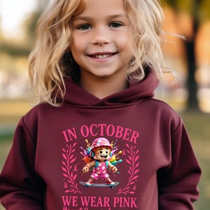 May include: A maroon hoodie with the text "IN OCTOBER WE WEAR PINK Breast Cancer Awareness". The graphic features a cartoon character on a skateboard, surrounded by pink accents and ribbons. The hoodie is worn by a child with blonde hair.