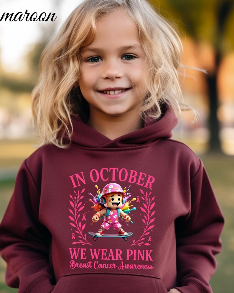 May include: A maroon hoodie with the text "IN OCTOBER WE WEAR PINK Breast Cancer Awareness". The graphic features a cartoon character on a skateboard, surrounded by pink accents and ribbons. The hoodie is worn by a child with blonde hair.
