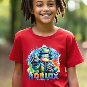 K&ouml;nnte beinhalten: Ein rotes T-Shirt mit einer farbenfrohen Grafik eines Roblox-Charakters mit Kopfh&ouml;rern, einem Controller und einem Friedenszeichen. Das Roblox-Logo ist in Regenbogenfarben dargestellt. Das T-Shirt wird von einer l&auml;chelnden Person getragen.