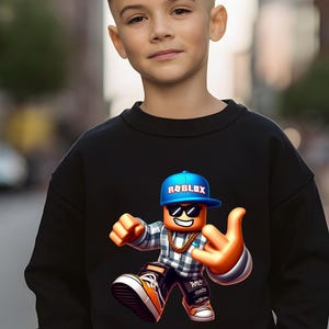 以下が含まれることがあります： 黒いスウェットシャツ。青い Roblox ロゴ入りのキャップ、サングラス、金のチェーンを身に着けた、カラフルな Roblox キャラクターが描かれています。キャラクターは手のジェスチャーをしています。スウェットシャツは若い人が着ています。