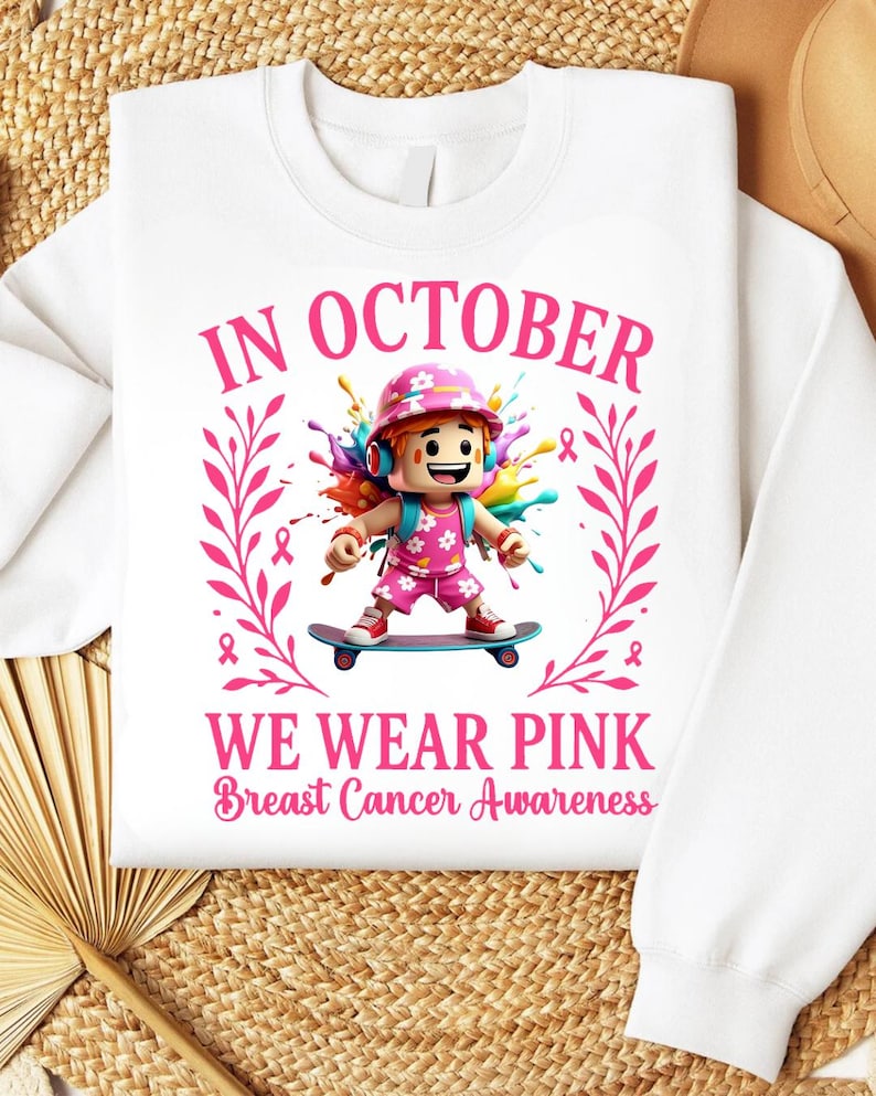 May include: White sweatshirt with a graphic promoting Breast Cancer Awareness. The design features a cartoon character on a skateboard, surrounded by pink ribbons and the text "In October We Wear Pink".