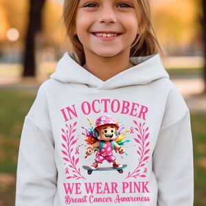 May include: White hoodie with the text "IN OCTOBER WE WEAR PINK Breast Cancer Awareness". The hoodie features a graphic of a character on a skateboard, surrounded by pink ribbons and floral elements. The word "white" is in the top left corner.